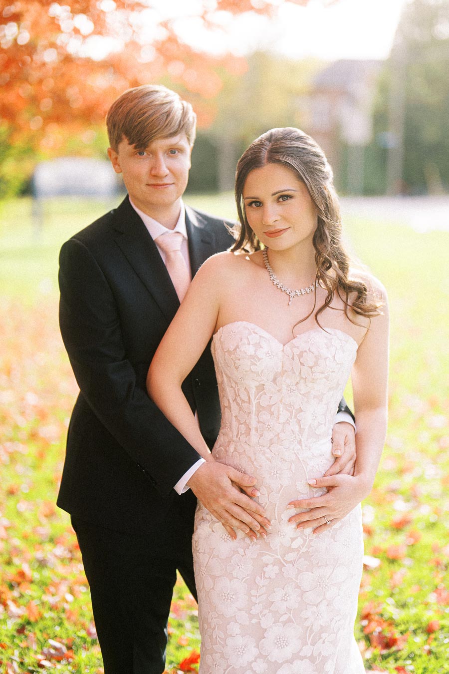 A bride and groom embracing in a sunlit park during autumn, with the bride in a strapless floral lace wedding gown and the