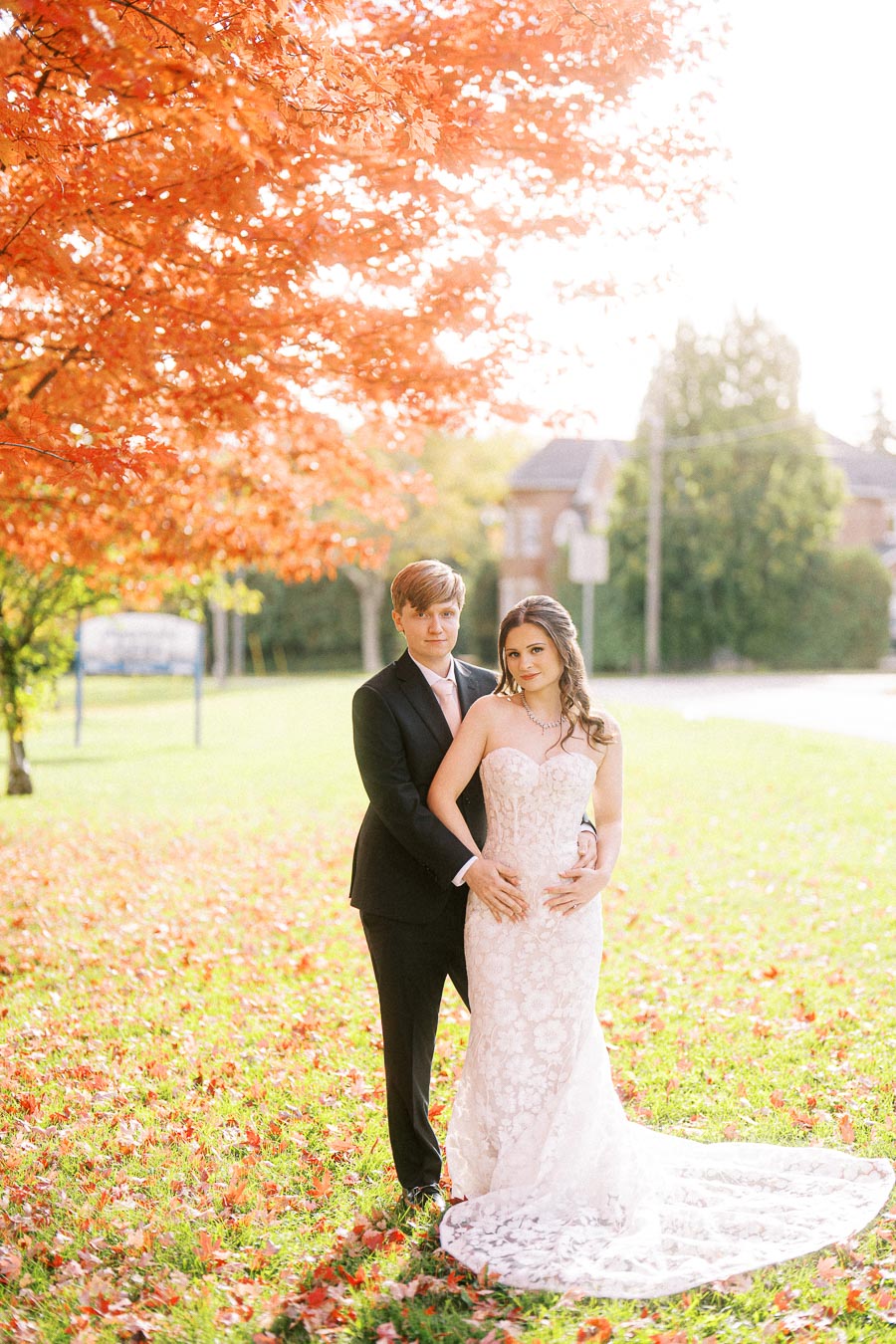 Bride and groom embracing under vibrant autumn leaves, wearing a lace wedding dress and a black suit in a picturesque