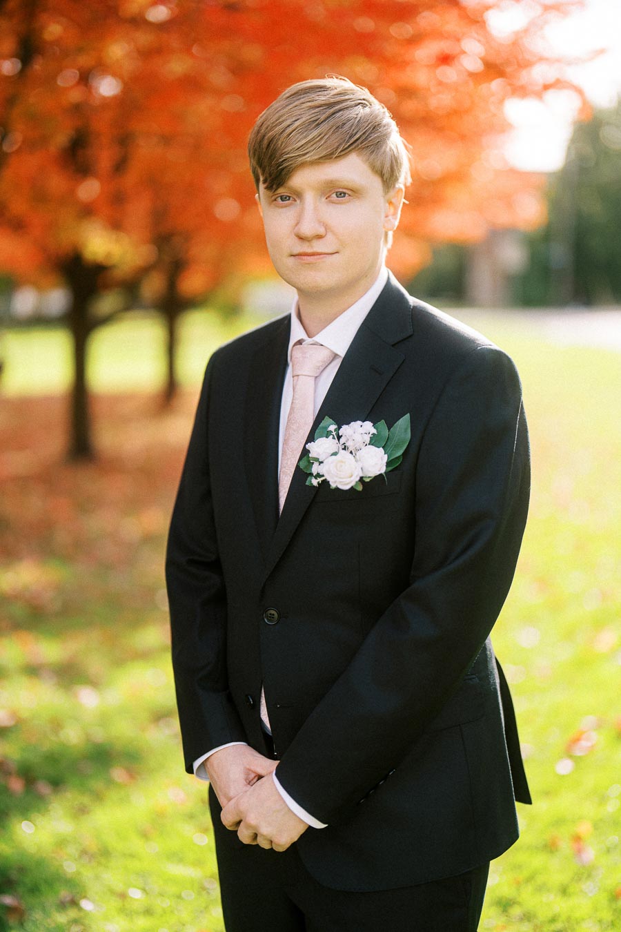 A young man in a black suit with a pink tie stands outdoors in front of vibrant red autumn foliage, with a boutonniere