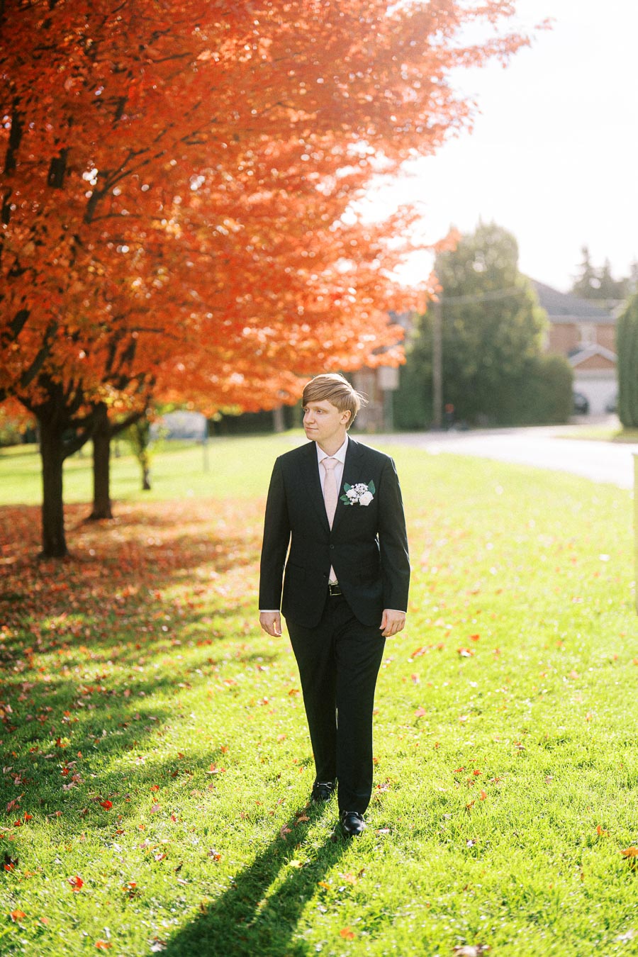 Man in suit walking on green grass under vibrant orange autumn leaves of a maple tree during a sunny day.