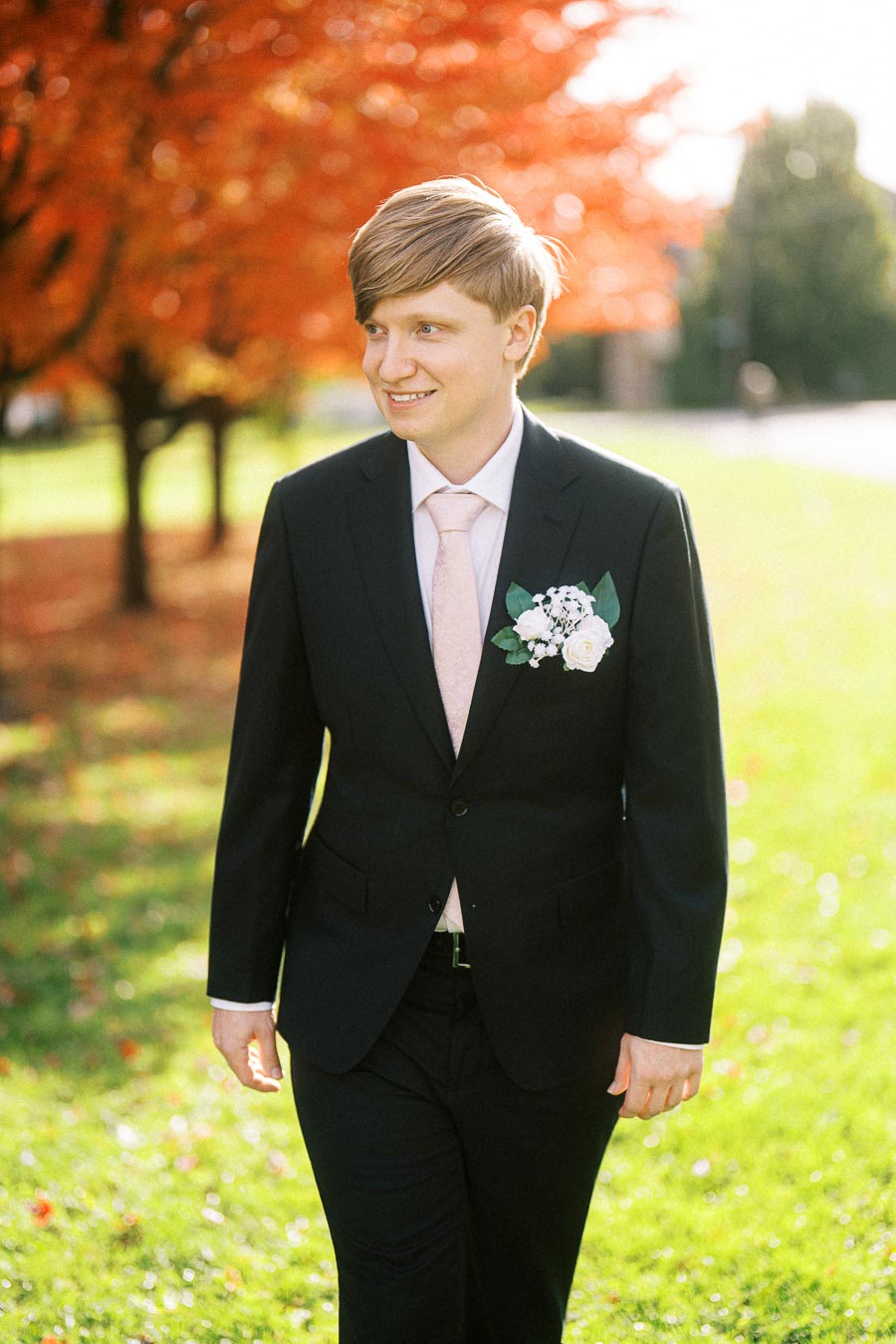 A person in a dark suit and pink tie walking outdoors in a park with vibrant autumn foliage and sunlight.