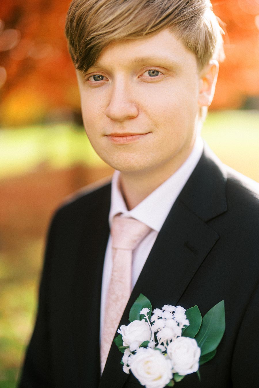 Young man in a black suit with a white floral boutonniere, standing outdoors with a blurred autumn background.