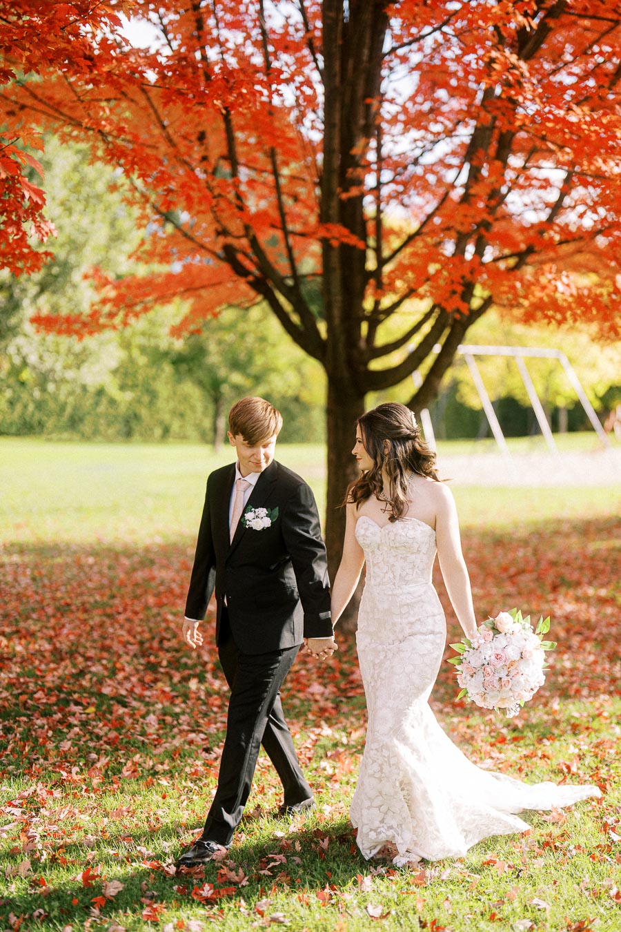 A bride and groom hold hands under a vibrant red tree during their outdoor autumn wedding, surrounded by fallen leaves.