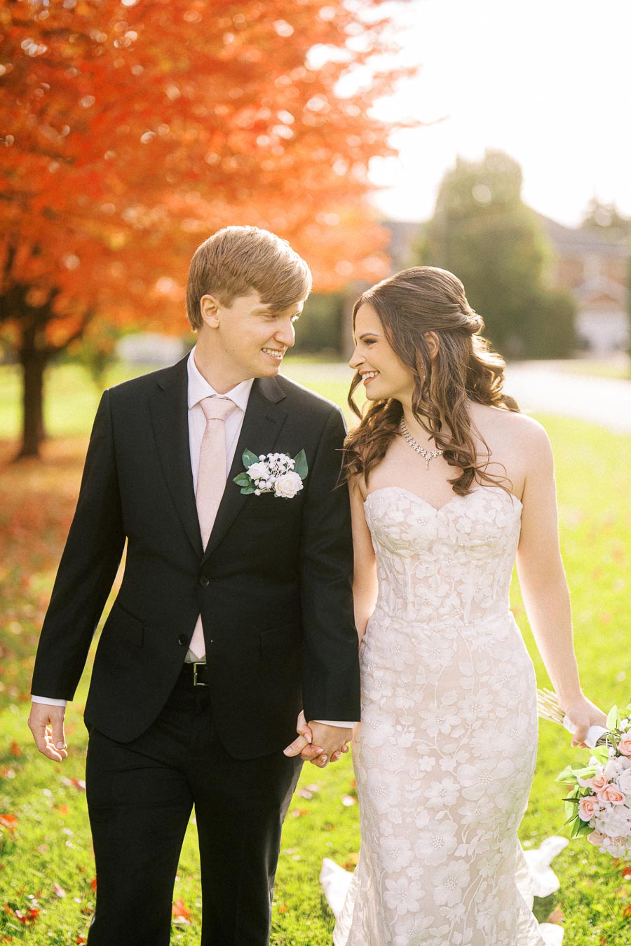 Smiling bride and groom holding hands in front of vibrant autumn foliage, dressed elegantly in a wedding gown and suit,