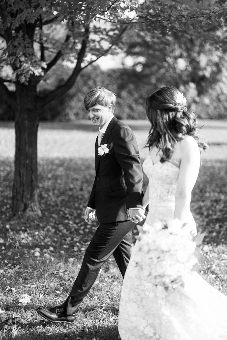 Black and white photo of a newlywed couple holding hands and walking outdoors, with the groom in a suit smiling and the