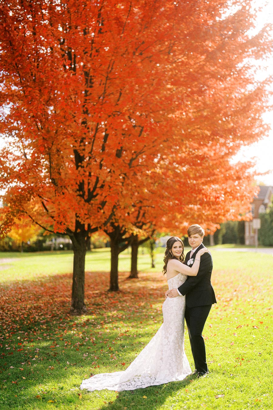 Wedding couple embracing under a vibrant orange maple tree in an autumn park, with the bride in a lace gown and the groom in