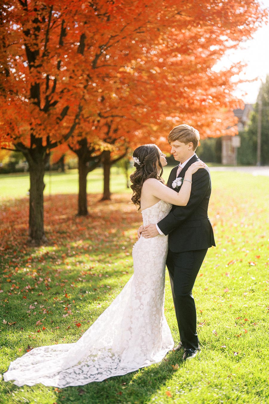 A bride and groom embrace under vibrant autumn trees, with the bride in a lace wedding dress and the groom in a black suit,