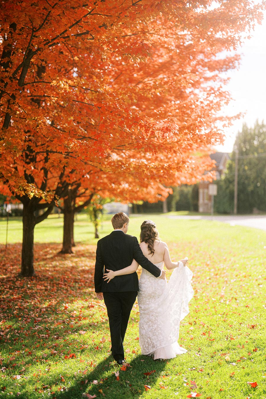 A couple in wedding attire walking hand in hand beneath vibrant orange autumn leaves on a sunny day.
