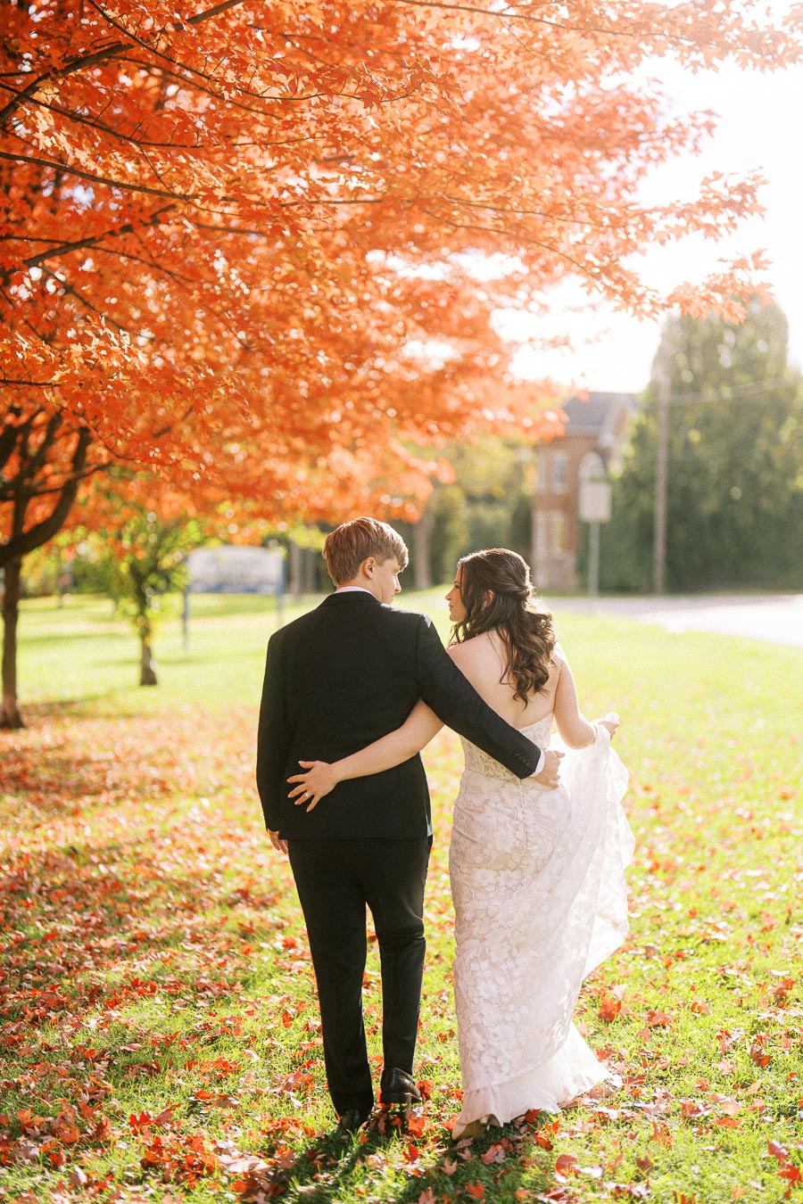 A couple in formal attire walks hand in hand under vibrant orange autumn trees, creating a romantic and picturesque scene
