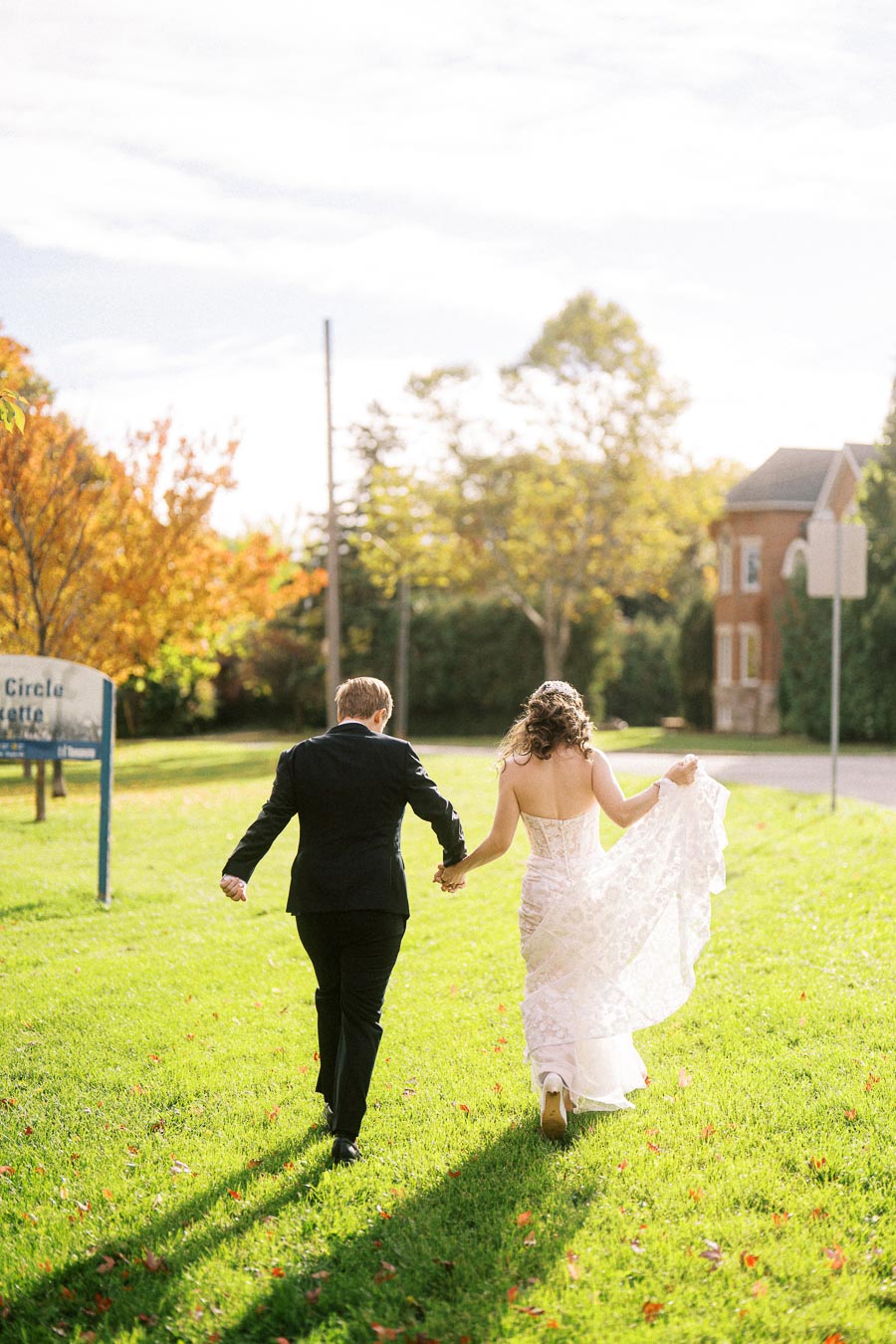 A bride and groom walking hand in hand on a sunny day, with autumn trees lining a lush green lawn, embodying a romantic