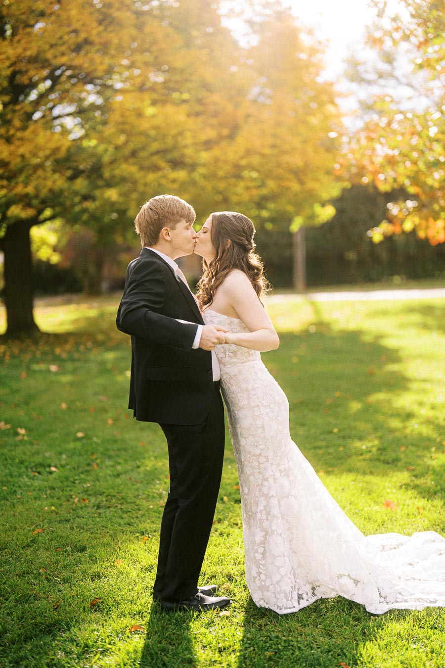Young couple in wedding attire sharing a romantic kiss in a sunlit park with autumn foliage.