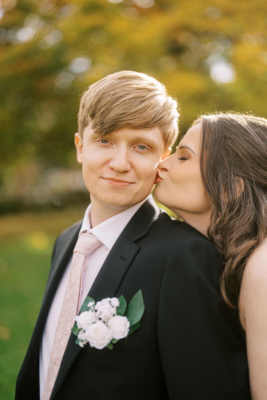 A bride kisses a smiling groom on the cheek during an outdoor wedding photo shoot. The groom is wearing a black suit with a