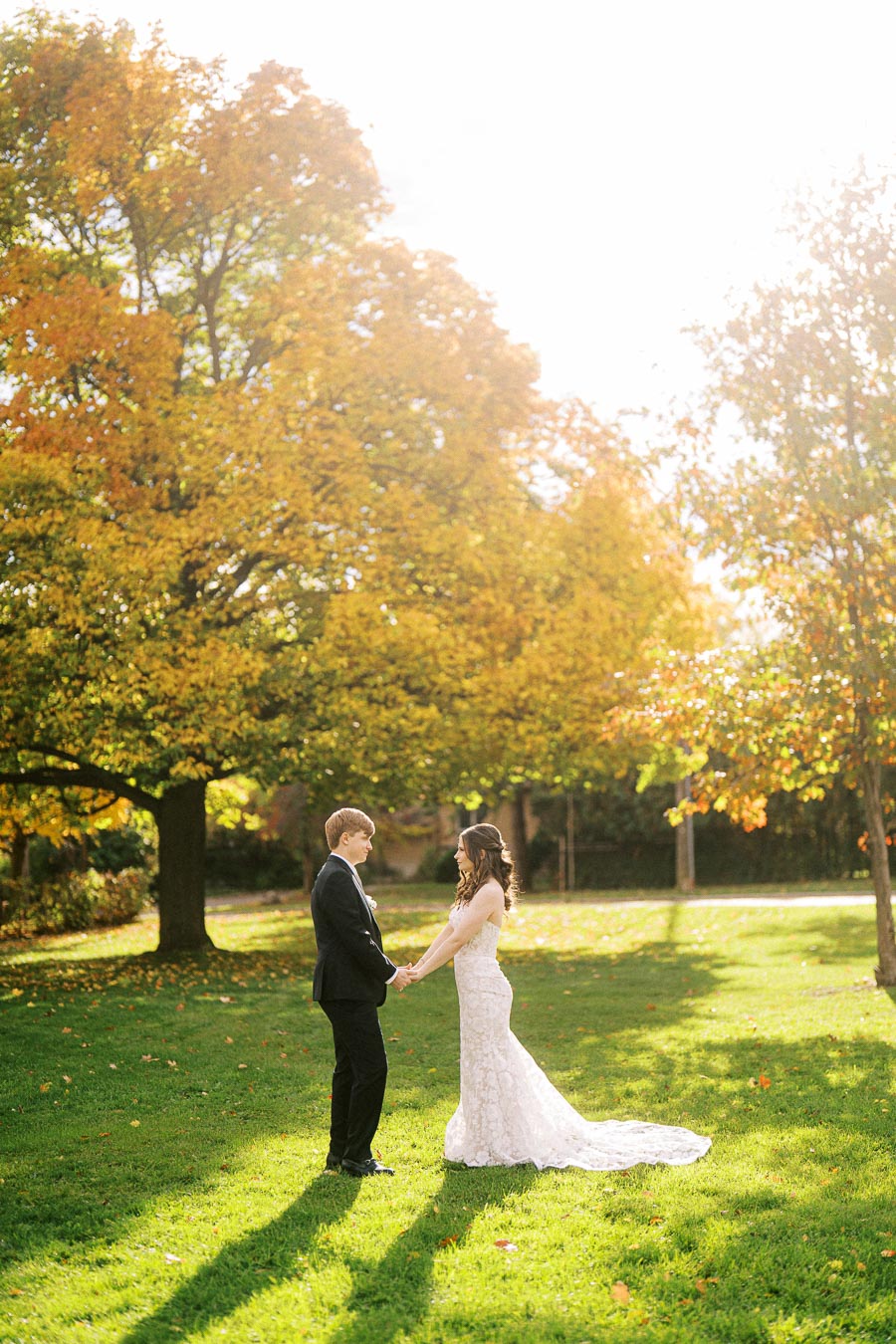 A bride and groom hold hands in a sunlit park, surrounded by vibrant autumn trees and green grass, perfect for a romantic