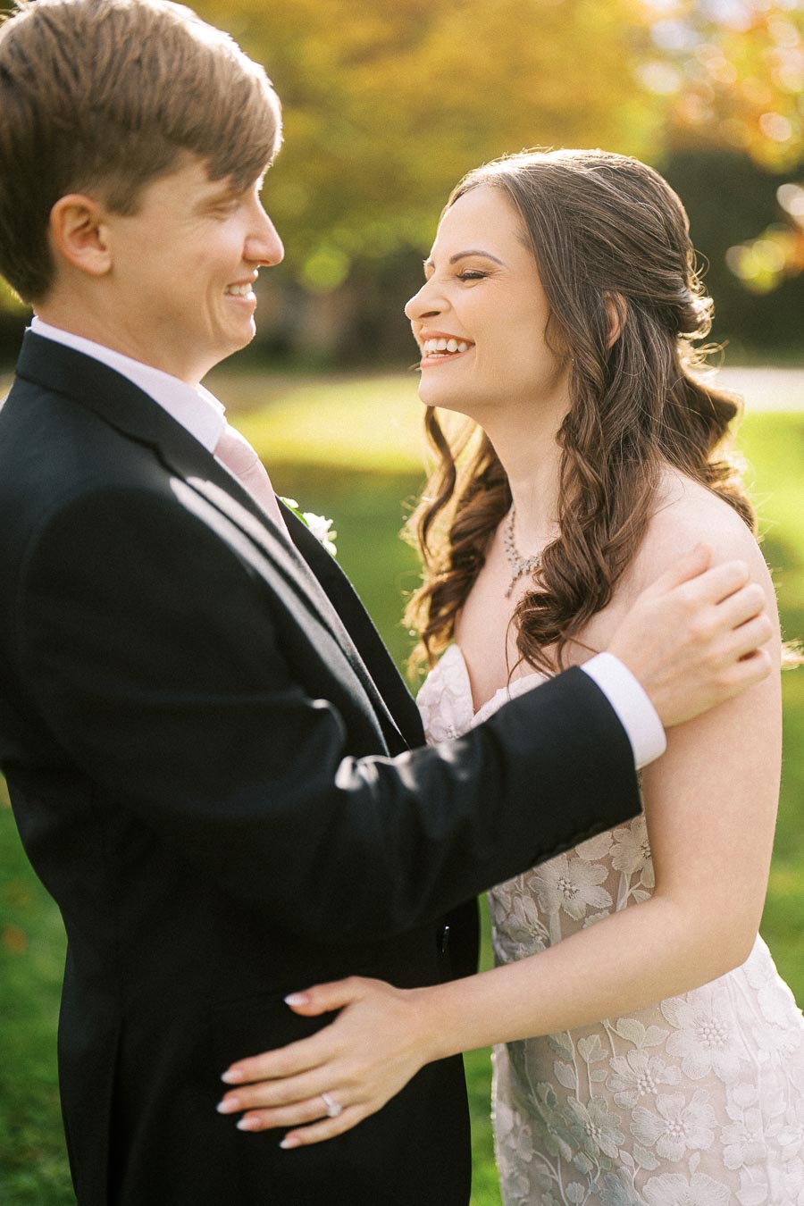 A joyful couple on their wedding day smiling at each other in a sunlit outdoor garden, with the bride in a lace wedding