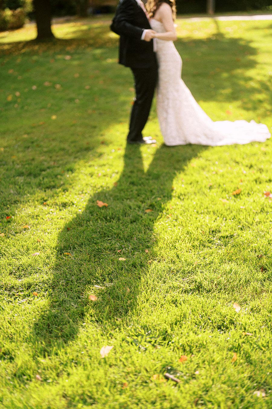 A bride and groom embrace on a sunny day, casting a long shadow on a lush green lawn, capturing a romantic moment outdoors.