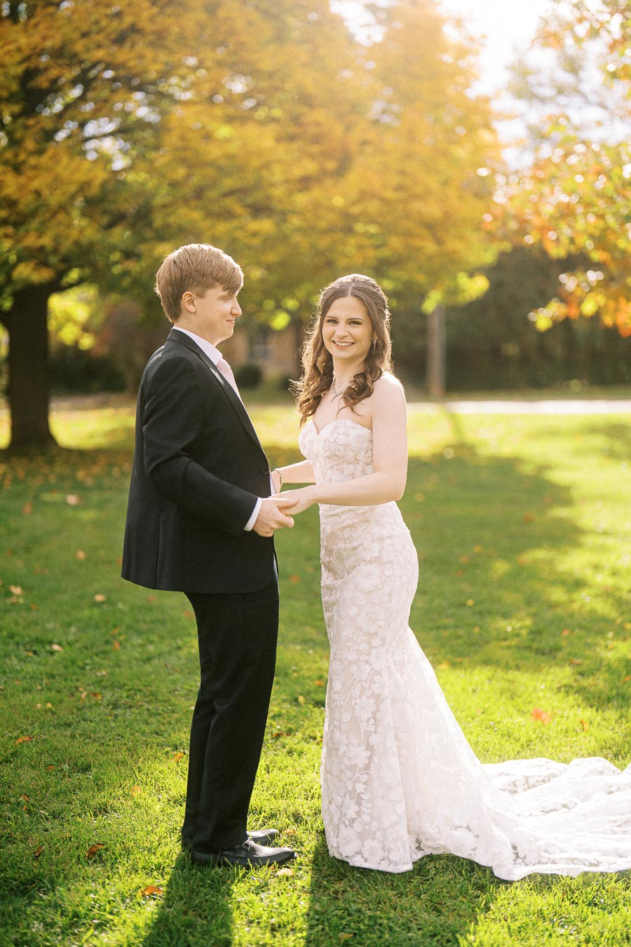 A newlywed couple smiling and holding hands in a sunlit park with autumn foliage, featuring the bride in a lace wedding