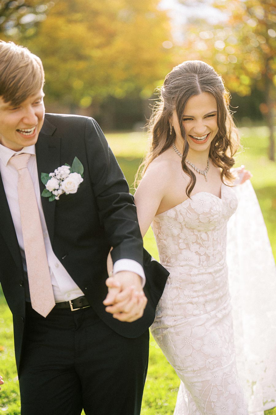 Happy couple holding hands and laughing at an outdoor wedding, with the bride in a lace dress and the groom in a suit with a