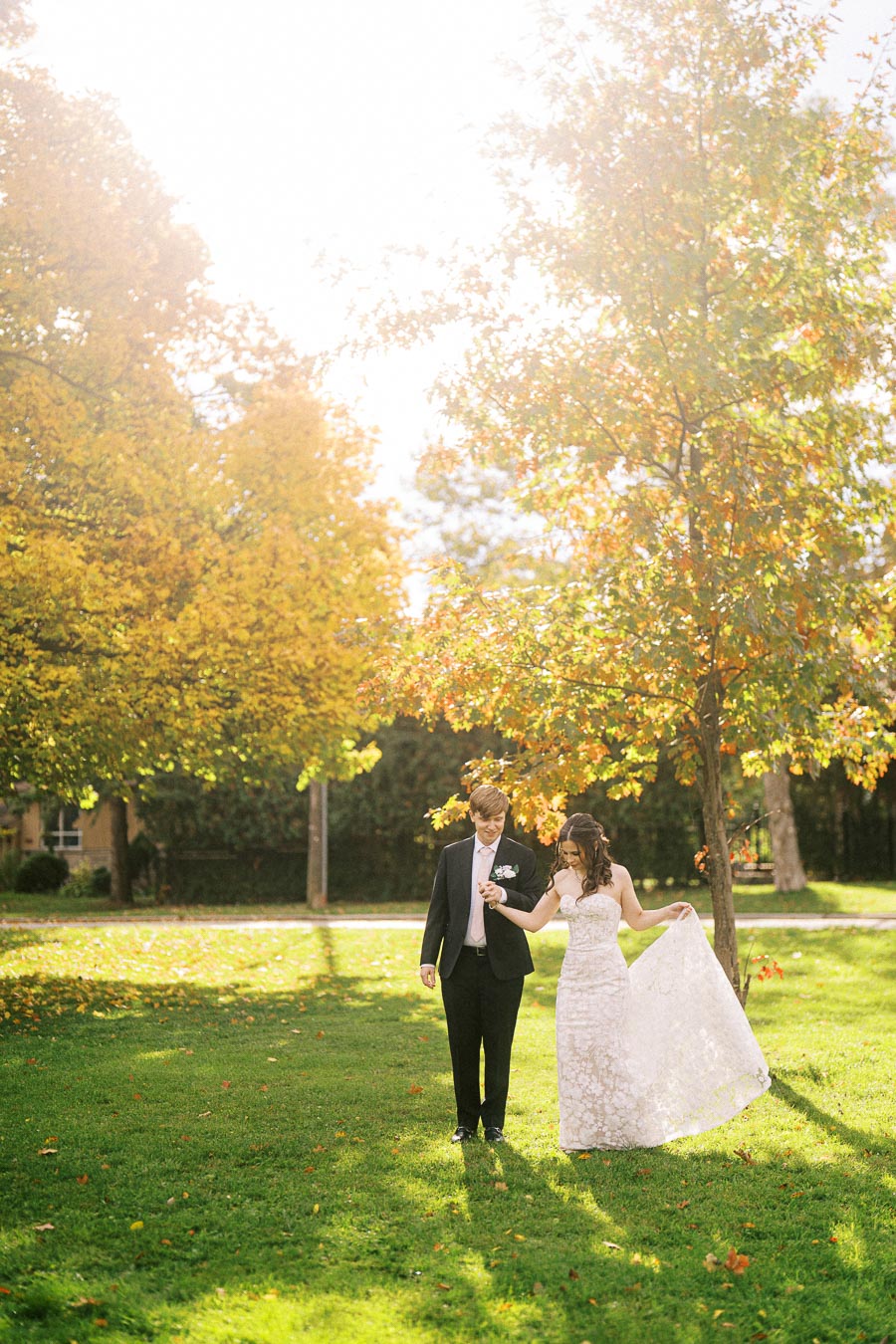 A bride and groom walking hand-in-hand on a lush green lawn during a sunny autumn afternoon, surrounded by colorful fall