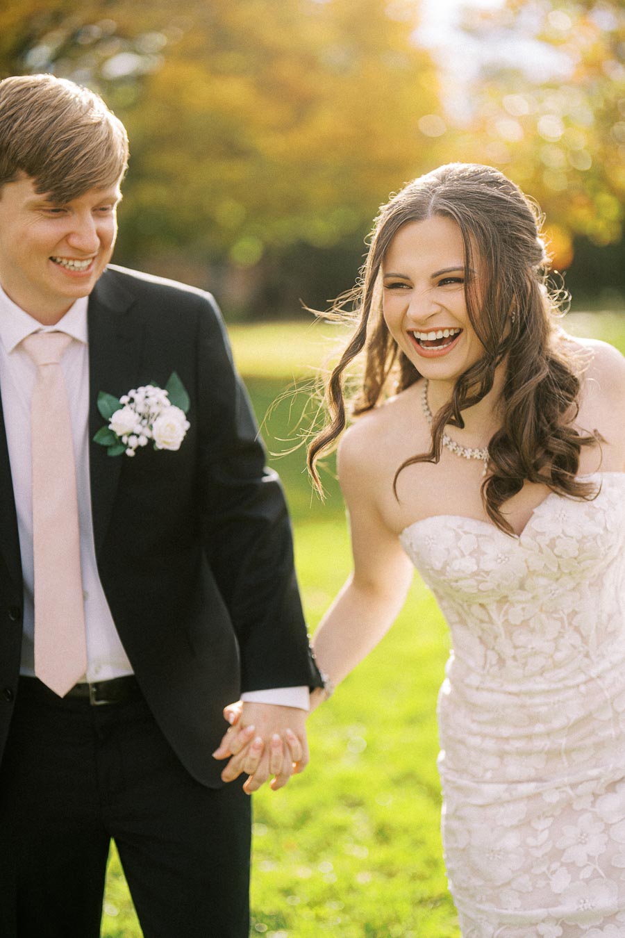A joyful bride and groom holding hands and smiling broadly in an outdoor setting with lush greenery and warm sunlight,