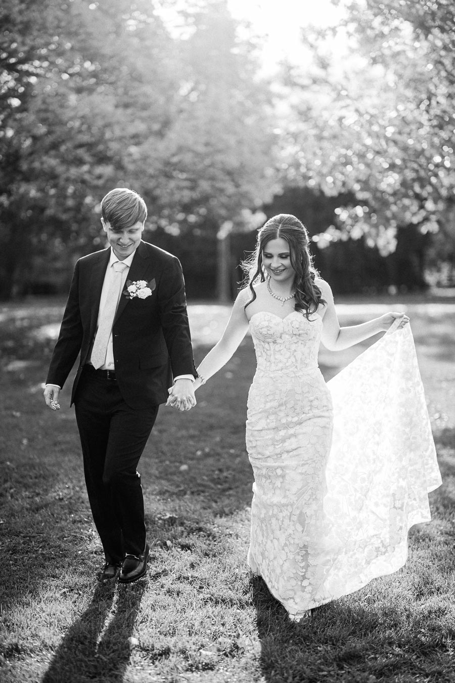 Elegant bride and groom holding hands while walking through a sunlit garden, capturing a timeless black and white wedding