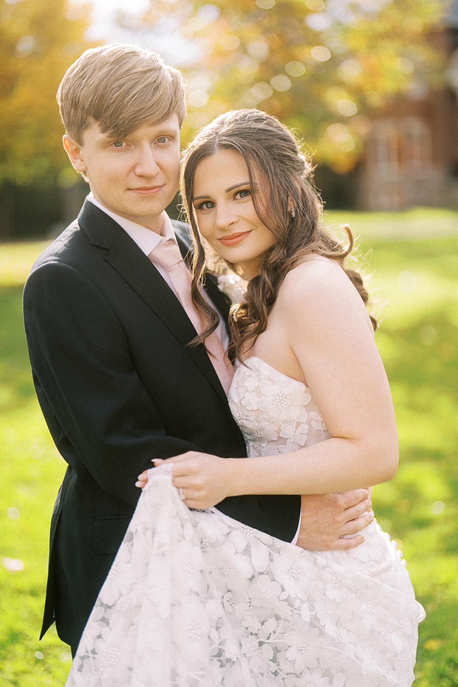 A couple in formal attire poses outdoors with a lush green background, the woman in a white floral gown embraces the man in