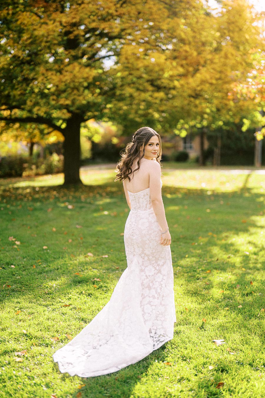 Bride in elegant lace wedding gown standing on green grass with autumn trees in the background, basking in