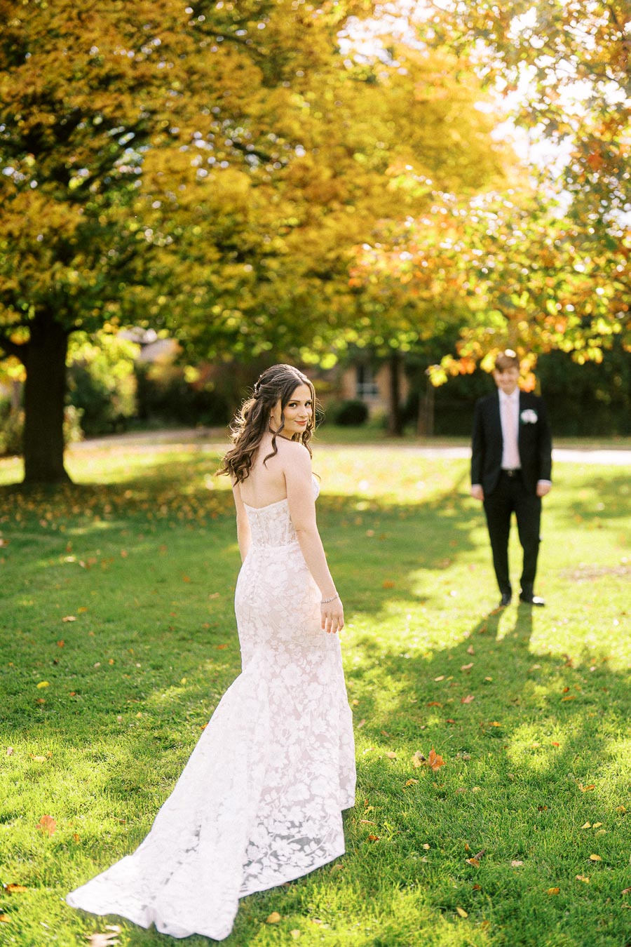 Bride in a white lace gown smiling at the camera, with a groom in a suit in the background, set in a sunny park with vibrant
