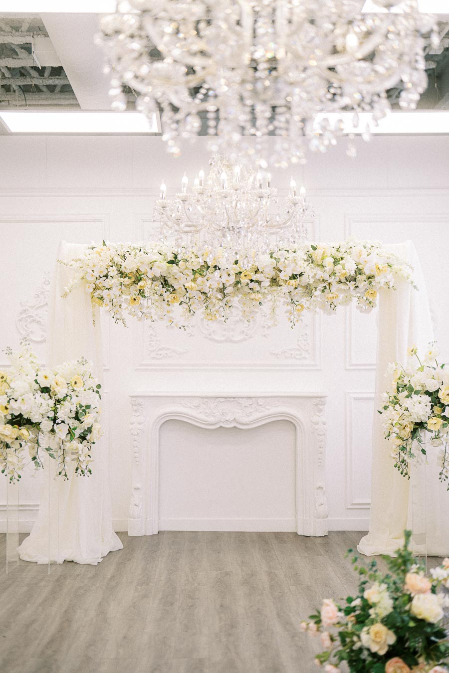Opulent wedding ceremony setup with an elegant chandelier, floral arch of white and yellow flowers, and classic white decor