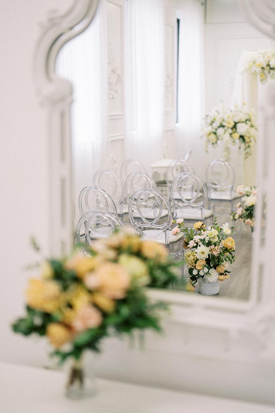Elegant wedding ceremony setup with clear acrylic chairs and soft yellow floral arrangements, reflected in an ornate mirror,
