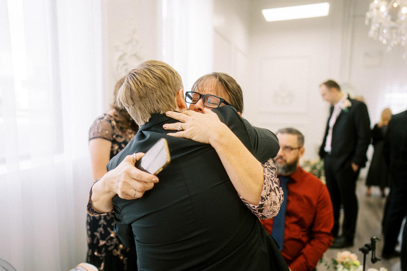 A heartfelt embrace between two people at a formal gathering, surrounded by elegantly dressed guests, emphasizing warmth and