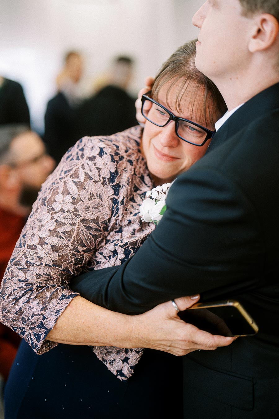A woman in a lace dress and glasses embracing a man in formal attire, holding a smartphone, at a social gathering.