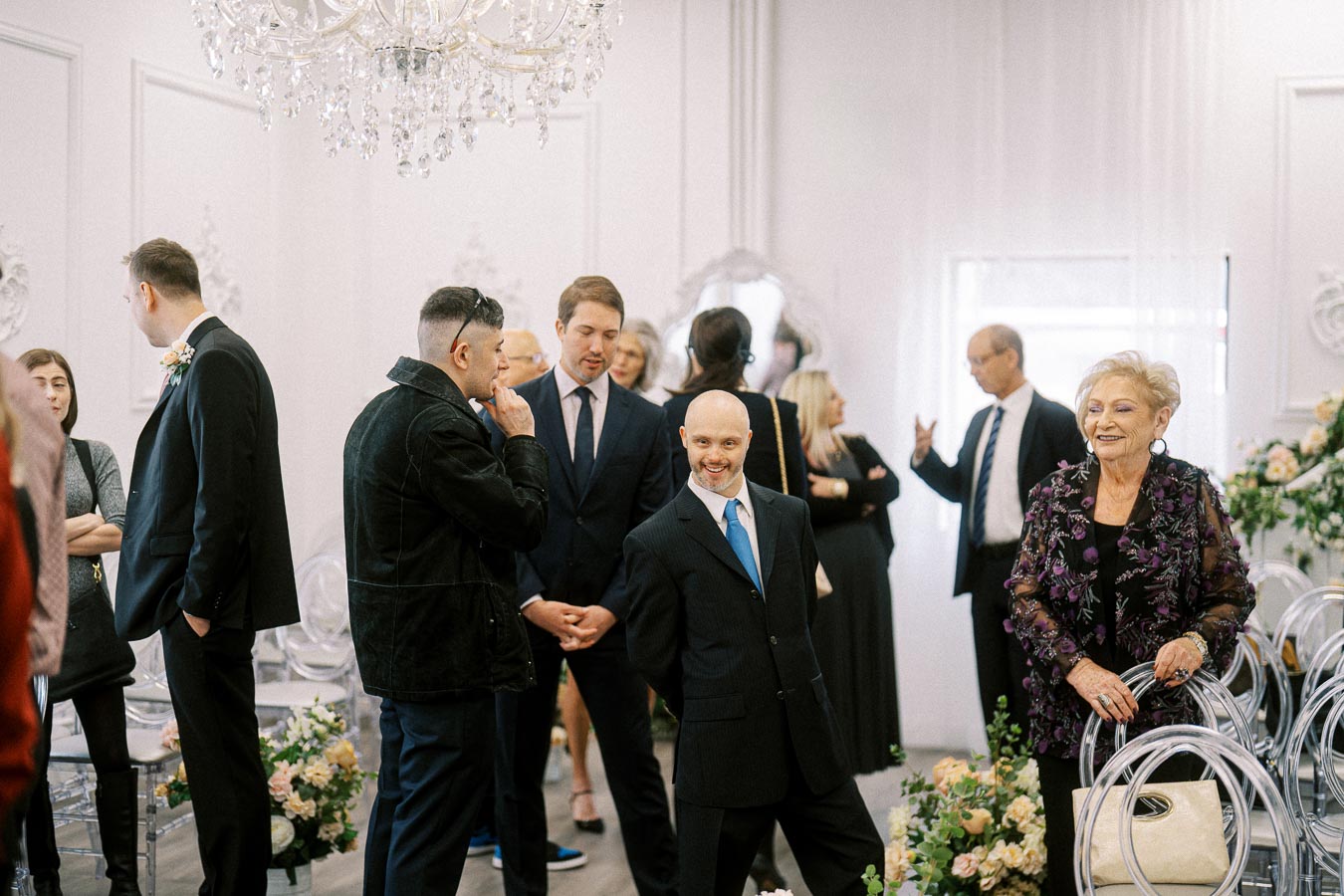 Group of people dressed in formal attire engaged in conversation at an elegant indoor event venue, adorned with chandeliers