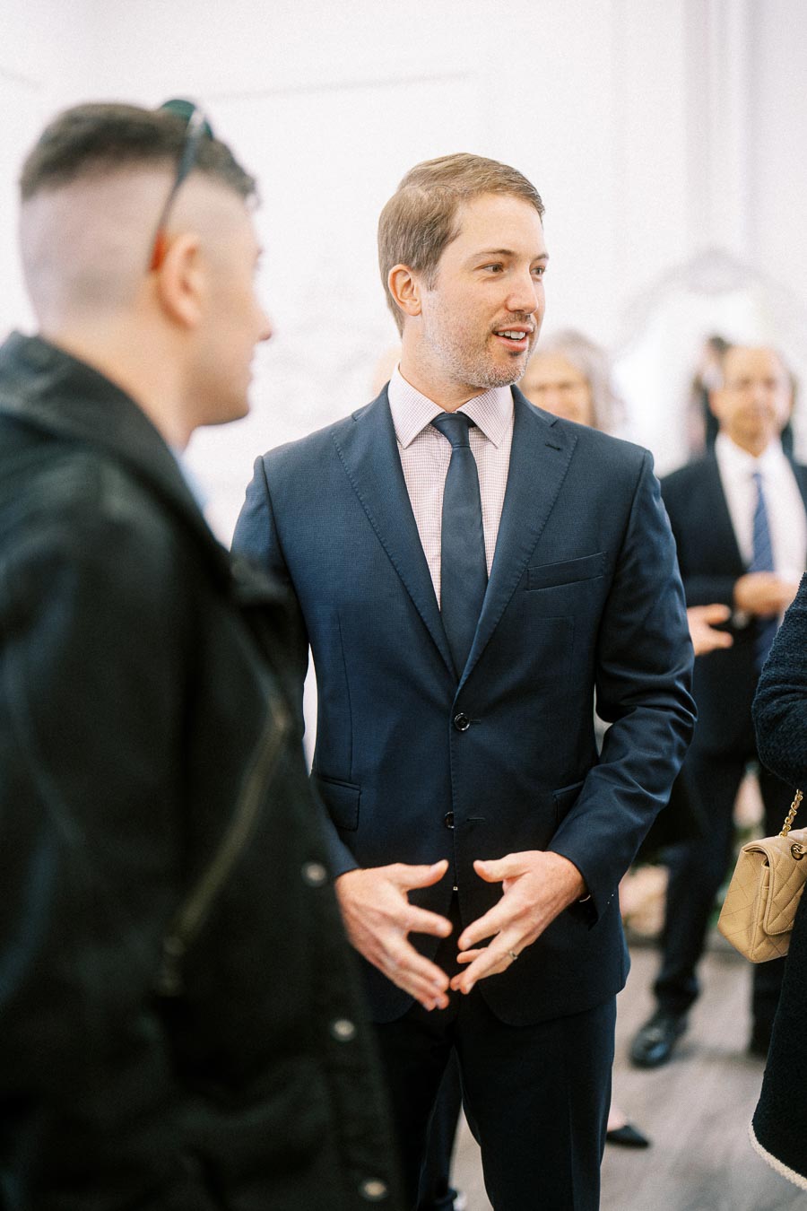 A professionally dressed man in a navy suit and tie engaging in conversation at a social or business event, surrounded by