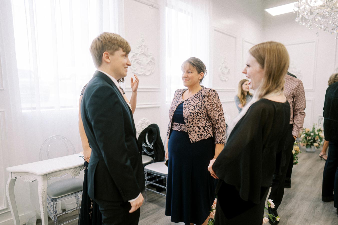 A group of people engaged in conversation at an elegant indoor gathering, featuring a man in a suit and women in formal