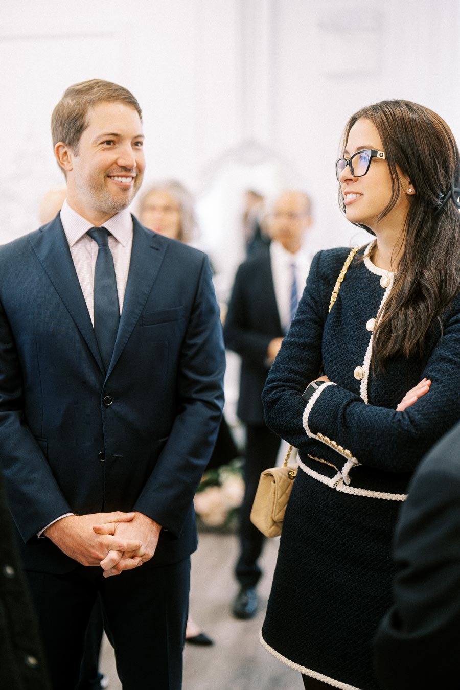 Two professionals in formal attire engaging in a conversation at a social event, with blurred attendees in the background.