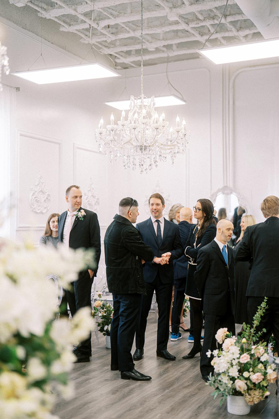 A group of people in formal attire mingling at a sophisticated indoor event venue with elegant chandeliers and floral