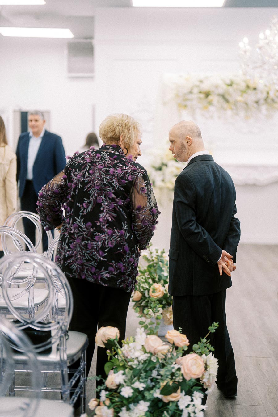 A well-dressed elderly woman and a bald man in a suit engaged in conversation at an elegant indoor event, surrounded by