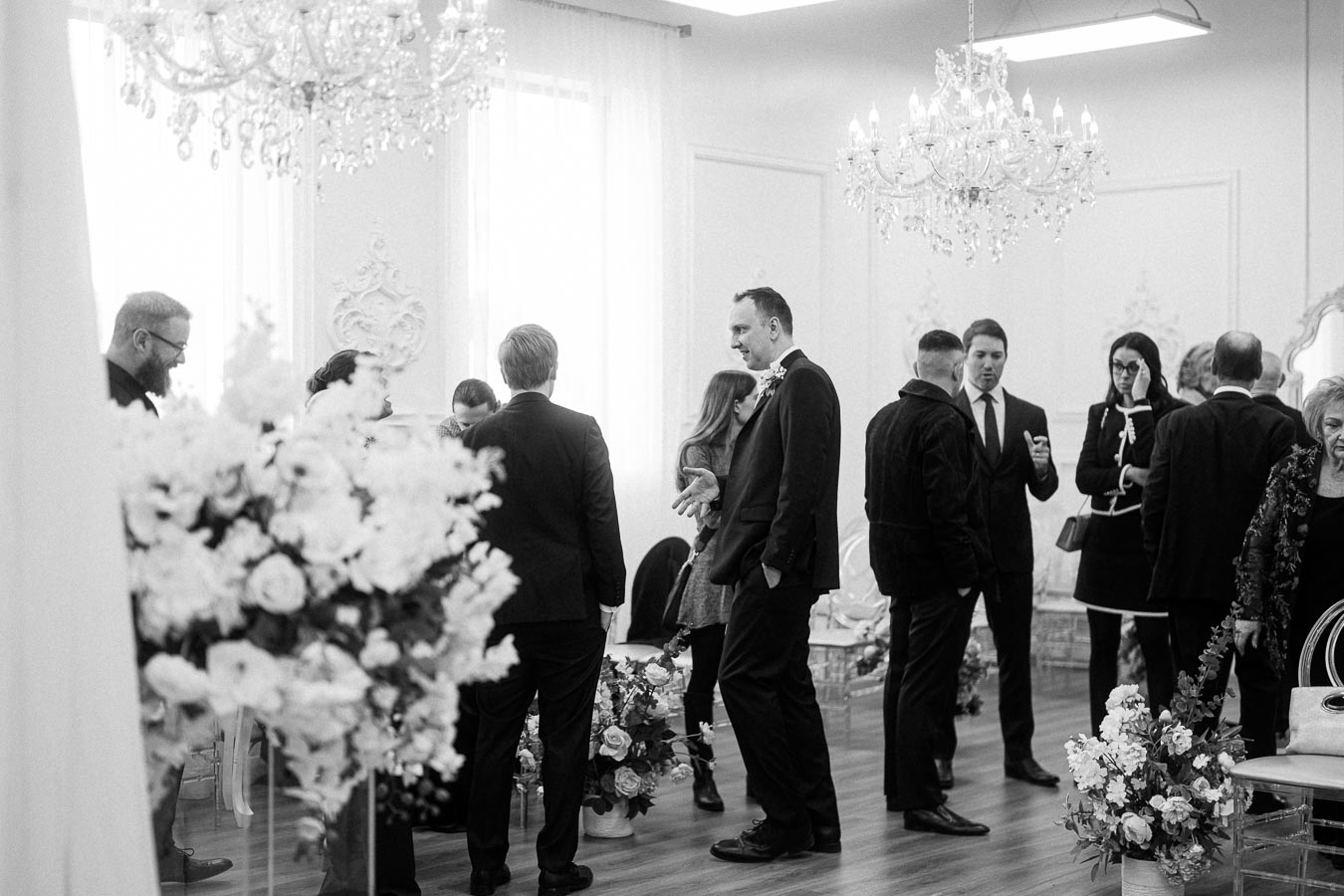 Black and white image of a formal gathering in a well-decorated room with chandeliers and floral arrangements. Several