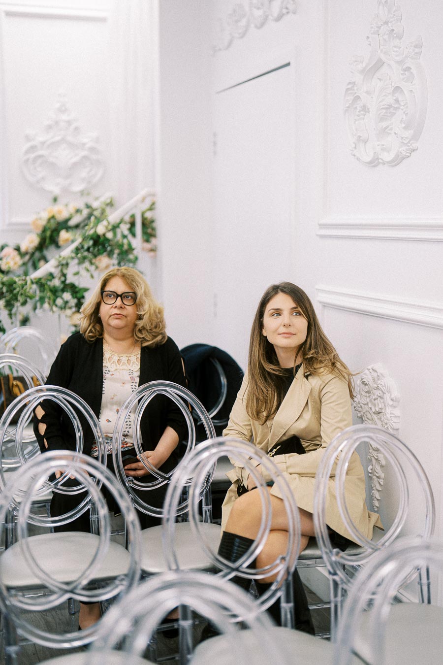 Two women sitting on transparent chairs in a bright, elegantly decorated room with floral arrangements and ornate wall