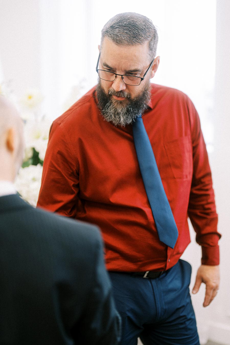 A man with a beard and glasses wearing a red shirt and blue tie, standing indoors, engaged in conversation.