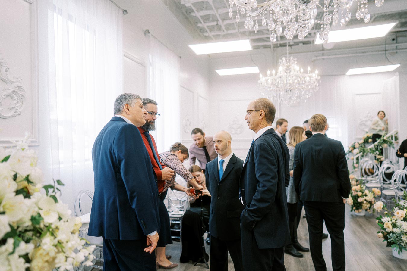 Group of people in formal attire engaged in conversation at an elegant indoor event with floral decorations and chandeliers.