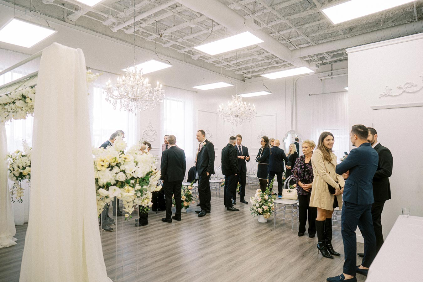 Group of people in formal attire socializing at an elegantly decorated indoor event space with floral arrangements and