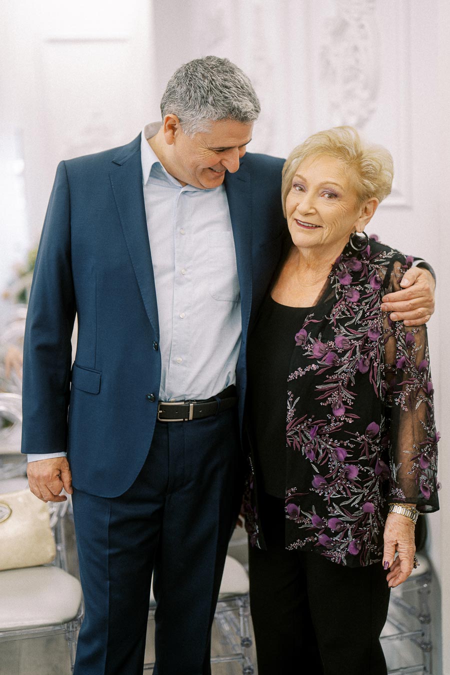 Smiling older woman in a floral jacket with a man in a blue suit embracing warmly in a brightly lit room.