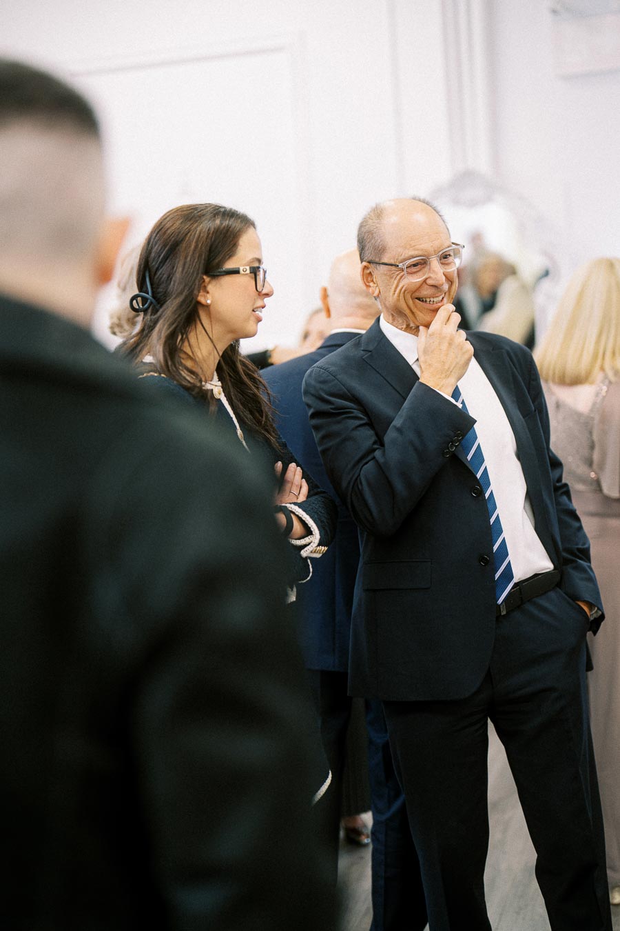 Two individuals in formal attire, a man with glasses wearing a suit and striped tie, and a woman with glasses in a black