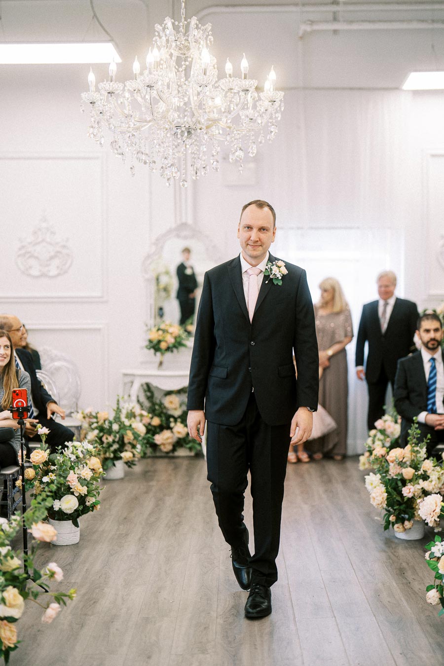 Groom walking down a flower-adorned aisle in an elegant indoor wedding ceremony, wearing a black suit and pink tie, under a