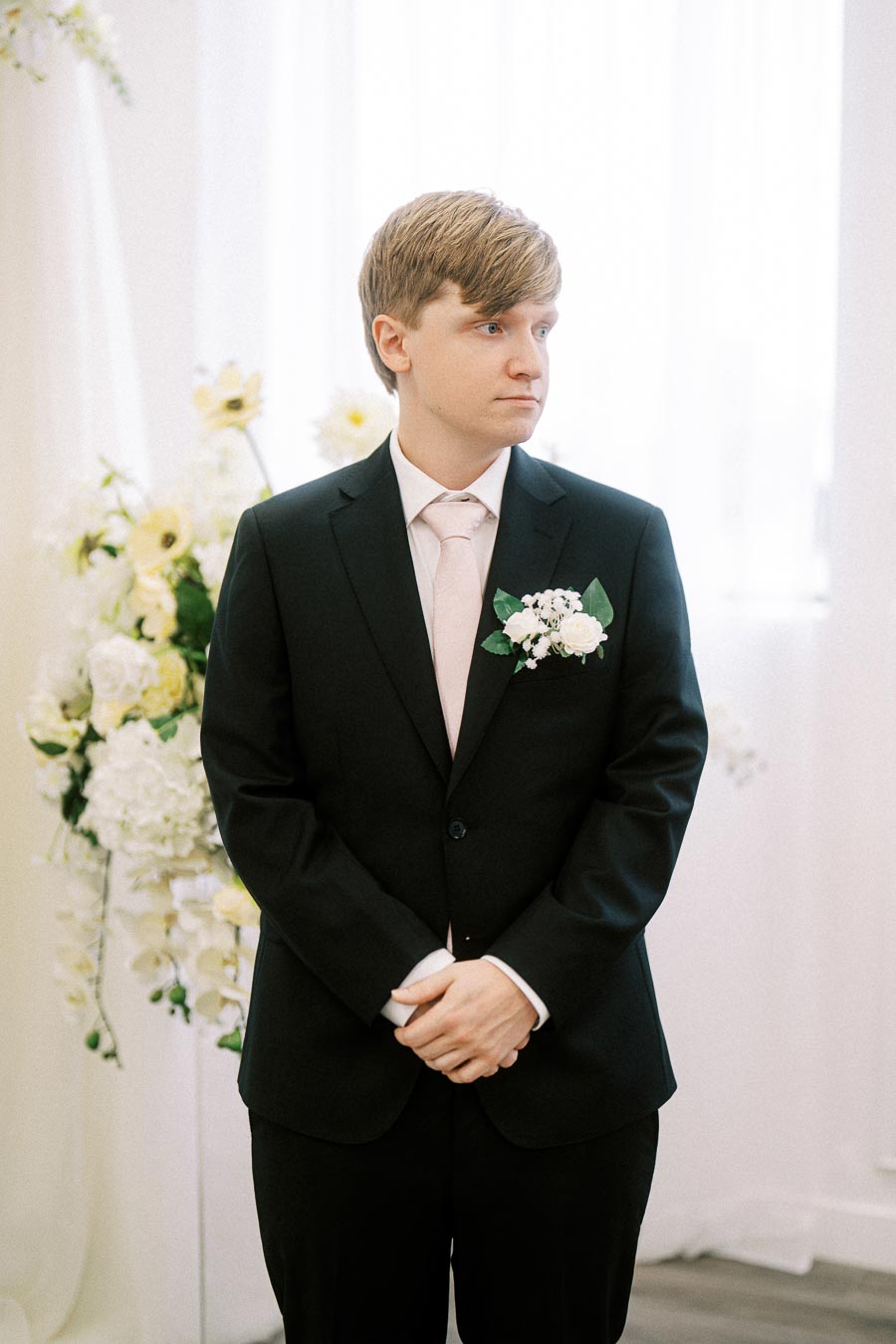 A person in a formal black suit and pink tie standing at an indoor wedding venue with white and yellow floral decorations in