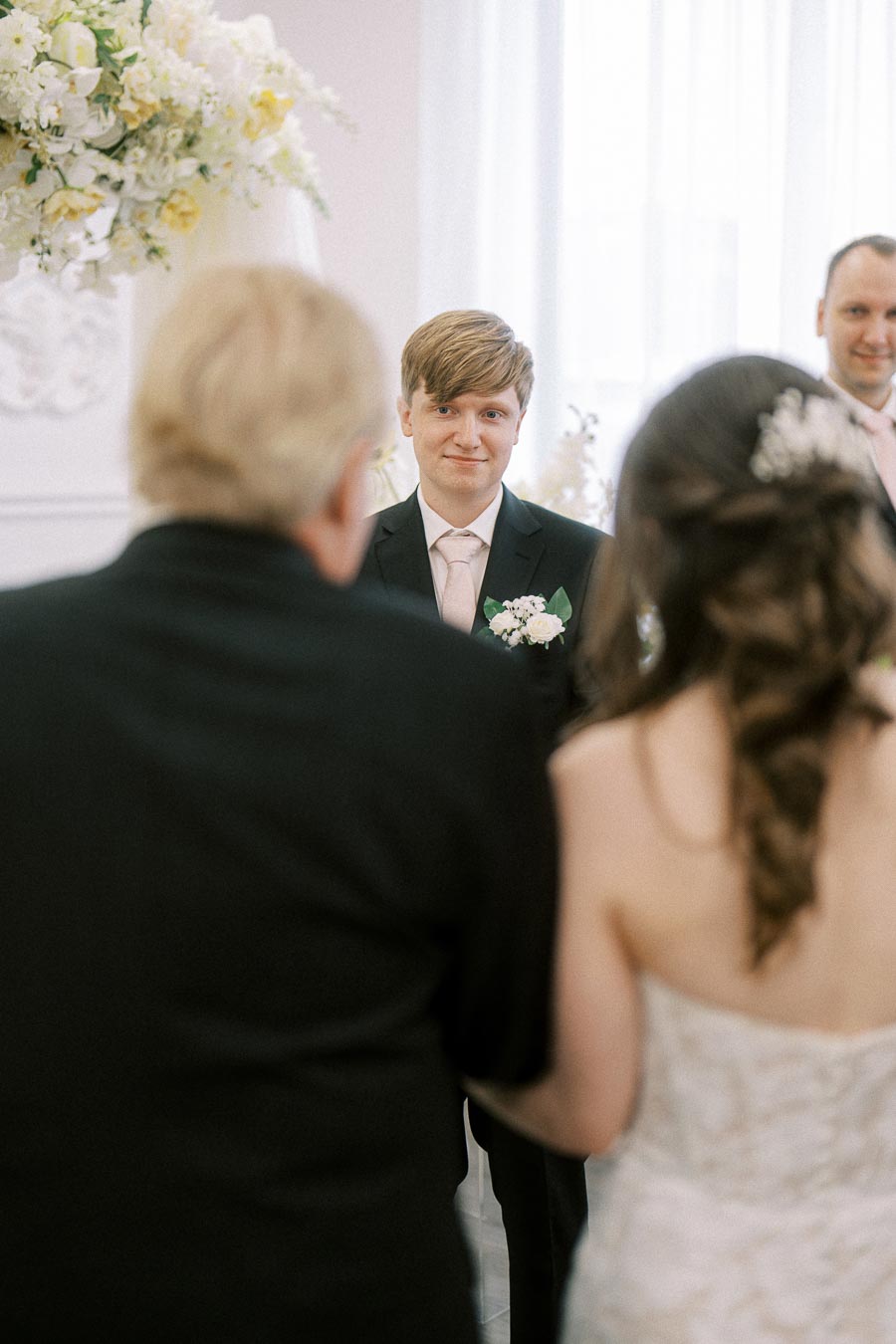 A groom in a black suit and pink tie smiling at his bride during a wedding ceremony, with floral decorations in the