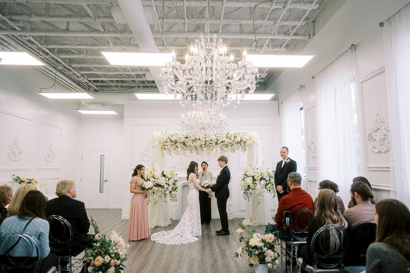 Elegant indoor wedding ceremony with a couple exchanging vows under a floral arch, surrounded by guests and bridal party in