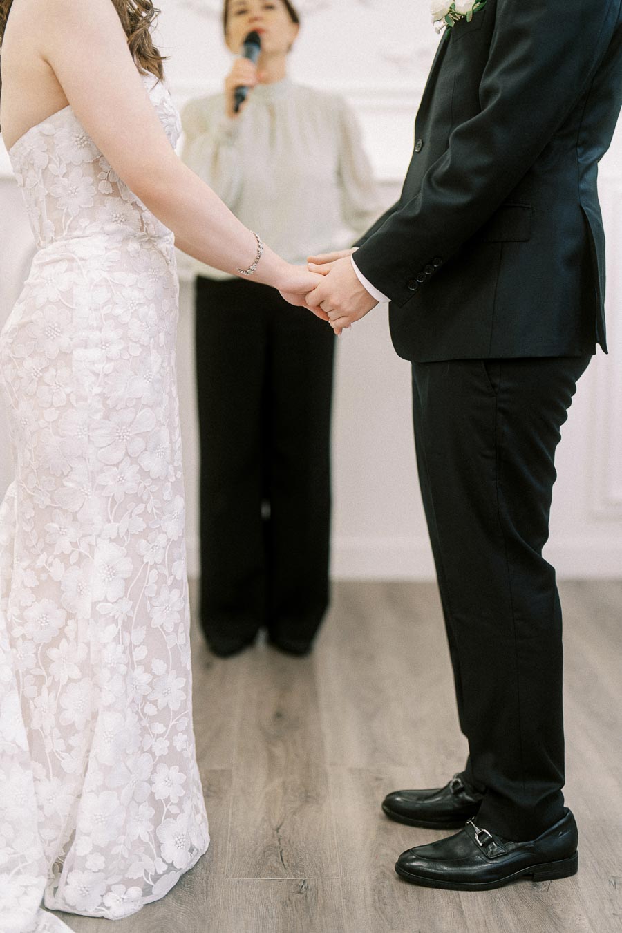 A bride and groom hold hands during a wedding ceremony, with an officiant speaking in the background.