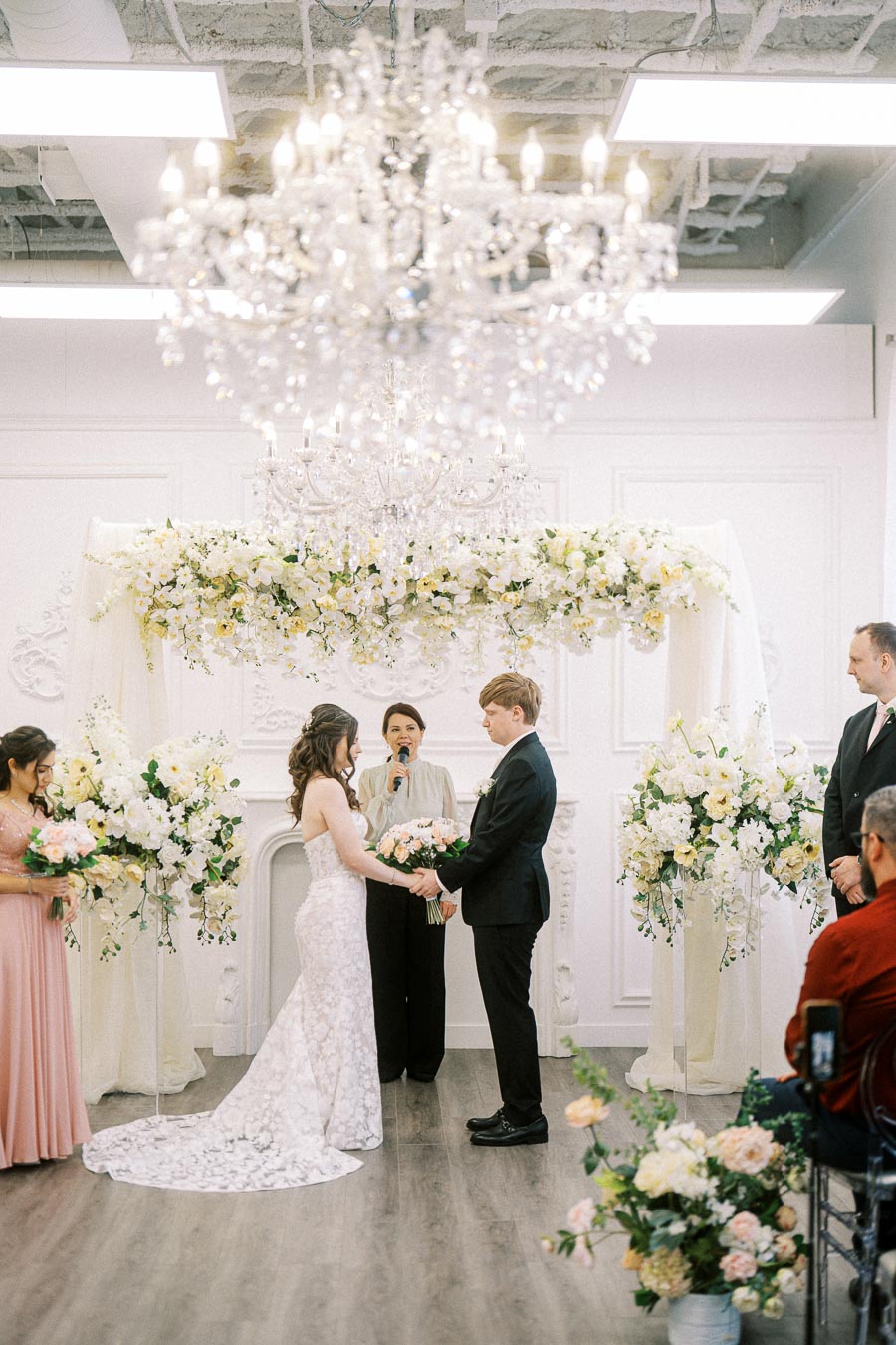 Elegant indoor wedding ceremony with couple exchanging vows under a floral arch, surrounded by floral arrangements and